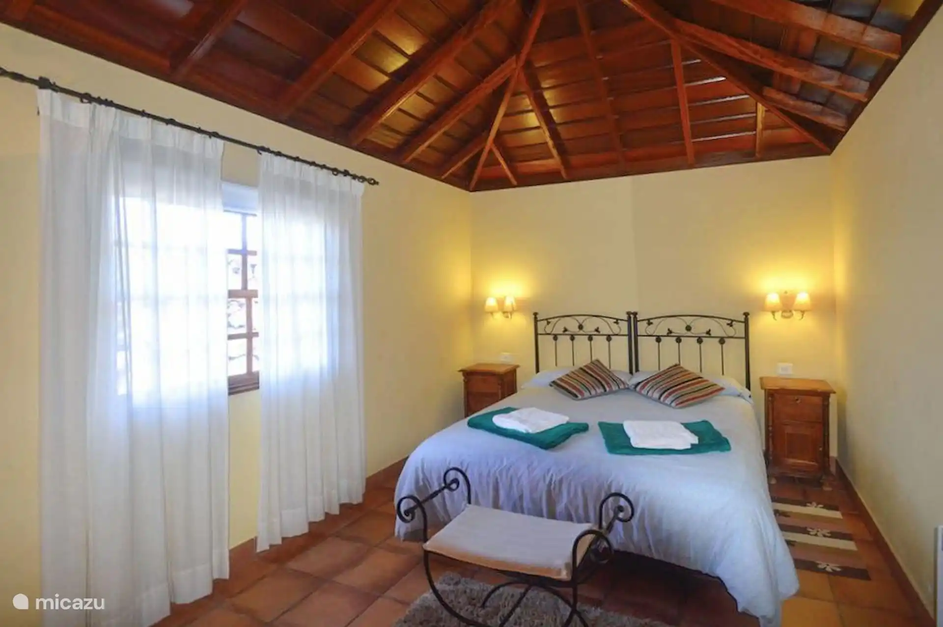 Bedroom with double bed, wrought iron headboard and wooden bedside tables; at the foot, a bench, and on top of it the wooden roof.