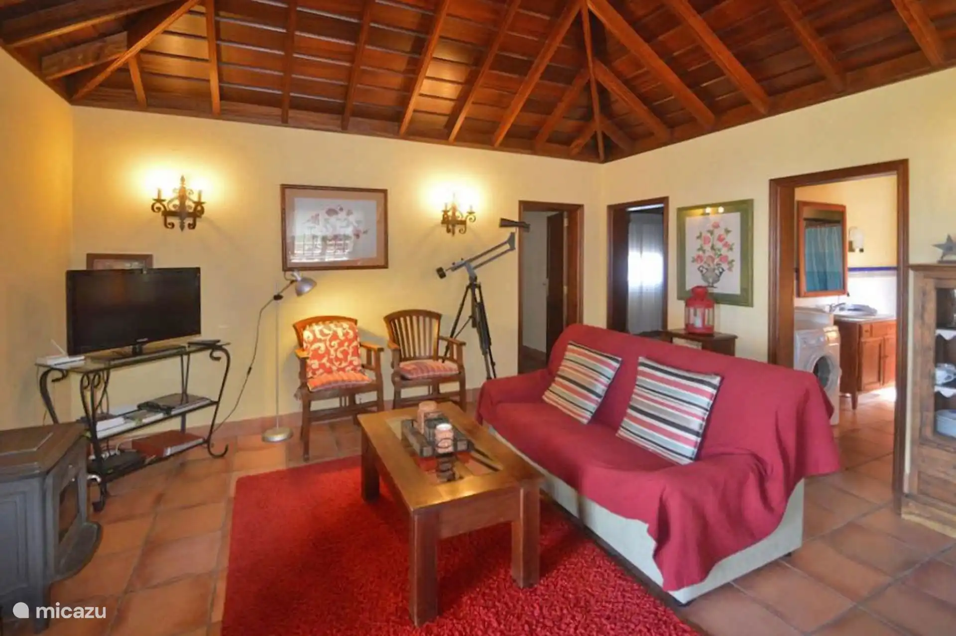Living room with sofa, two armchairs and television under the gabled wooden roof; on one side, the wood-burning stove and a telescope.