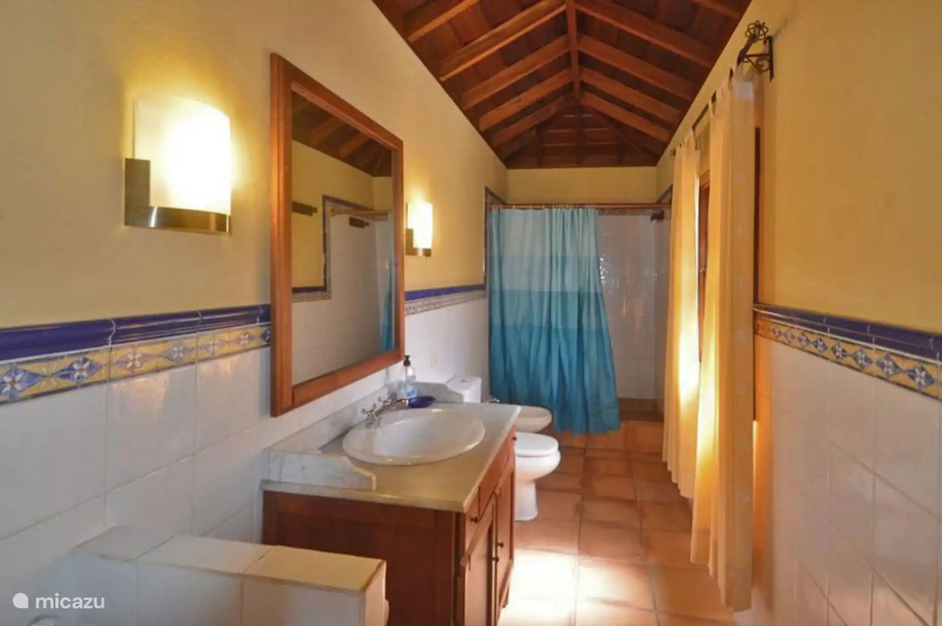 Elongated bathroom with sink on wooden furniture, mirror, toilet and shower with curtain; White tile with blue border.