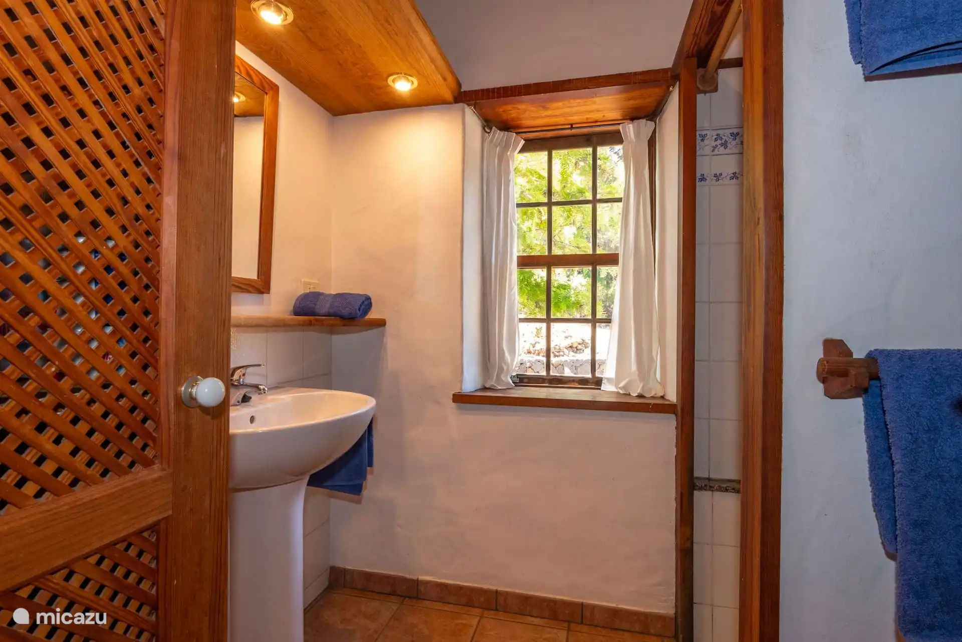 Badkamer met vrijstaande wastafel, spiegel en betegelde douche; Het houten raam kijkt uit op de tuin.