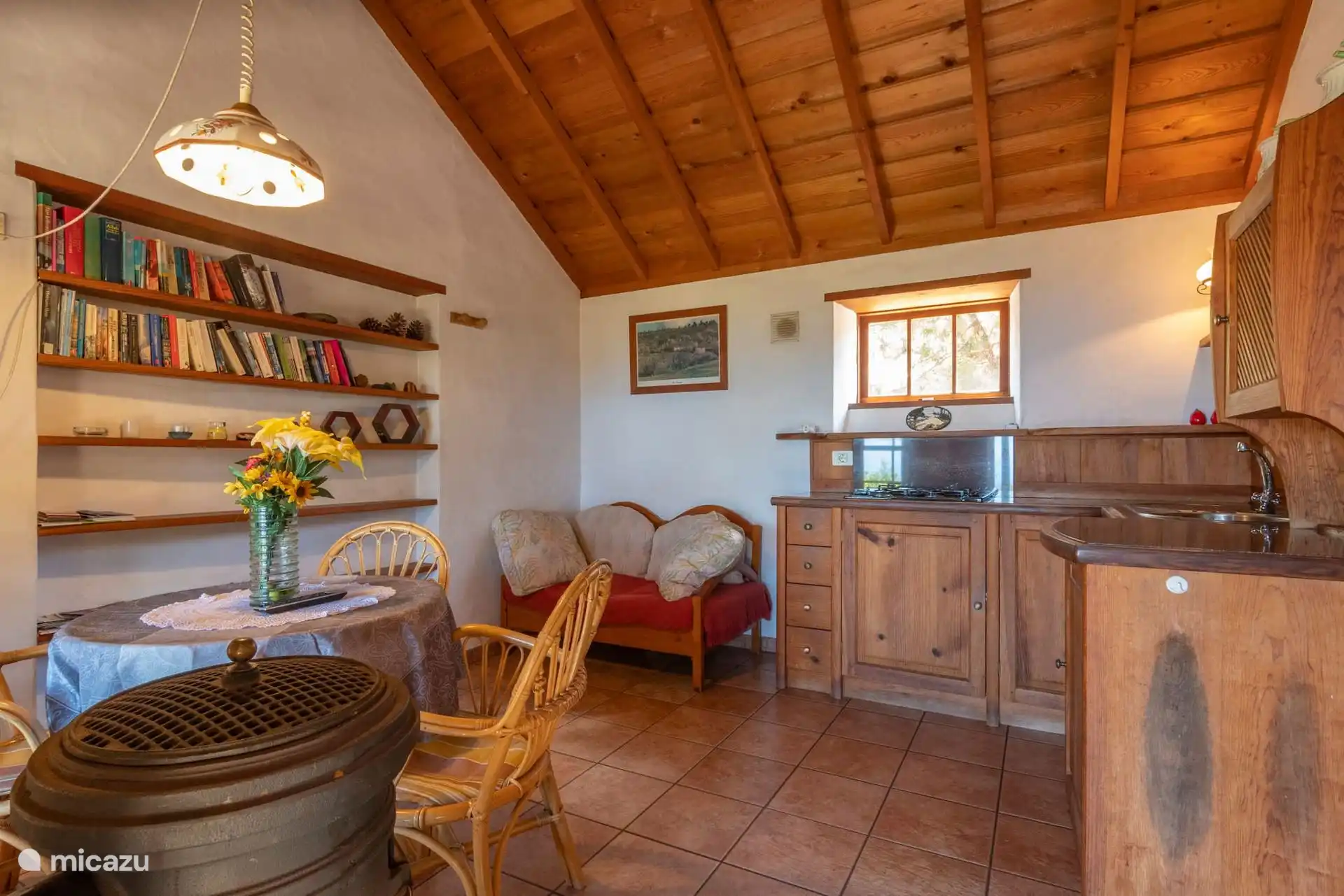 Keuken, eetkamer onder het houten zadeldak, met een ronde tafel met rieten stoelen, een bank en boekenkasten; De keuken, achterin achter het aanrecht.