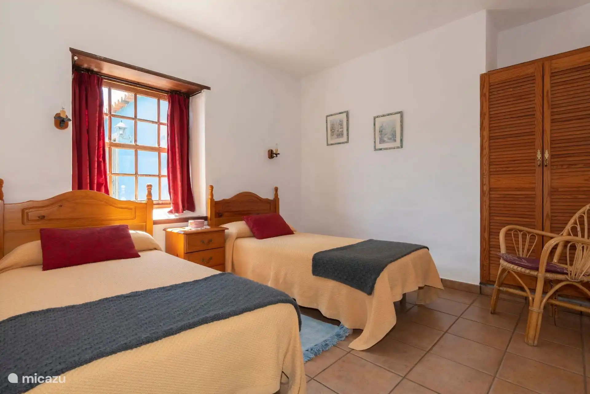 Bedroom with two single beds, slatted wardrobe and wicker armchair; The blue façade peeks out of the window.