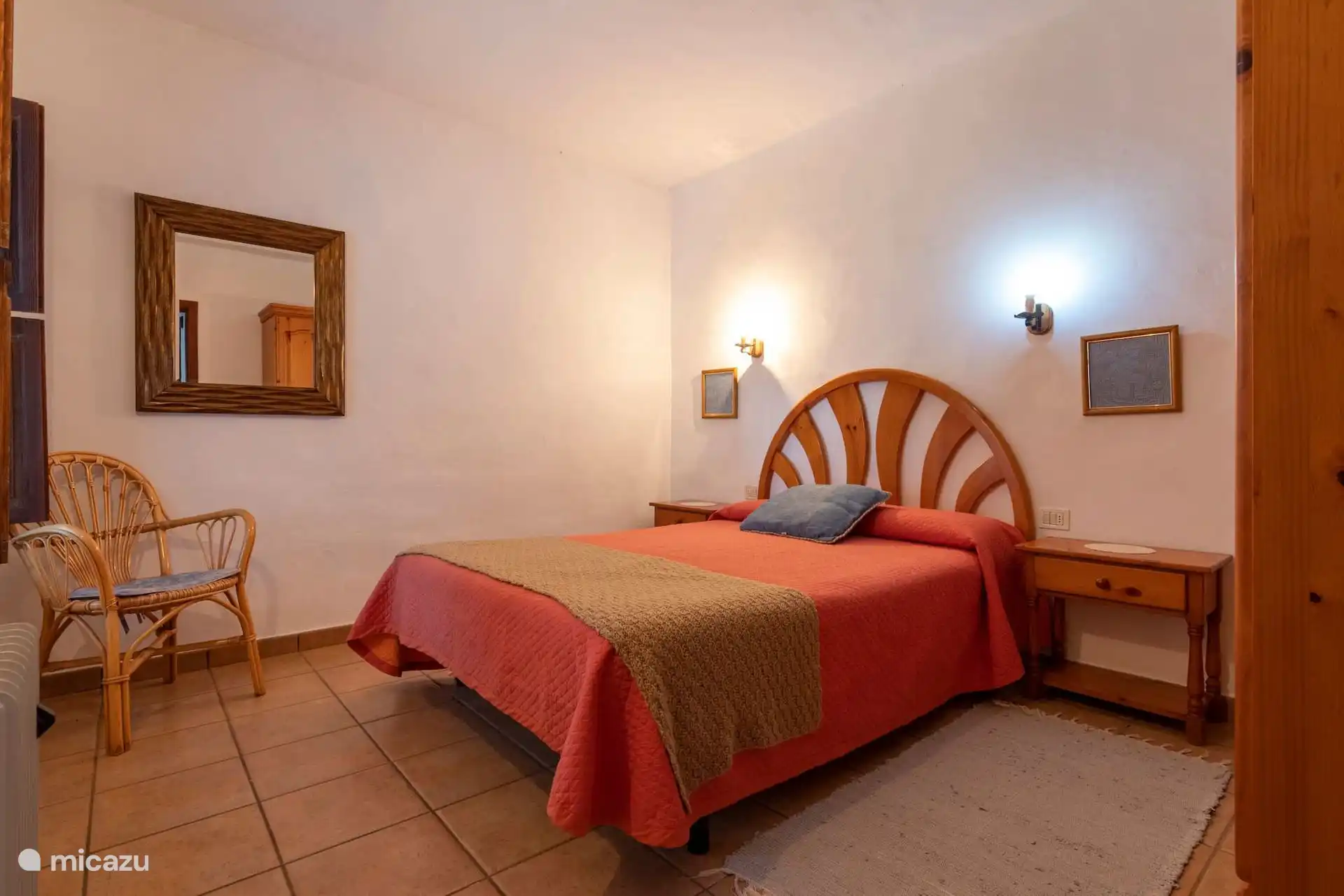 The same bedroom from the door, with mirror, wicker armchair and bedside tables on both sides of the bed.