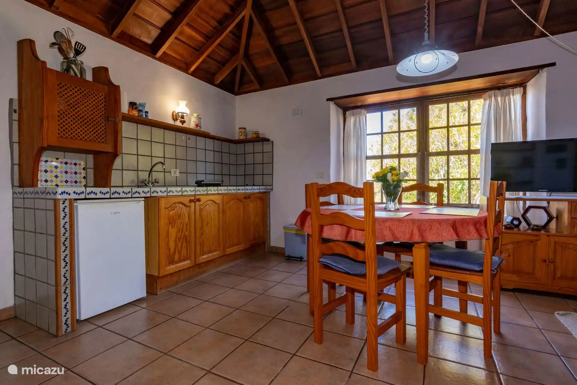 Kitchenette with sink, fridge, wooden cupboards and tiled worktop; next to it, the table by the window.