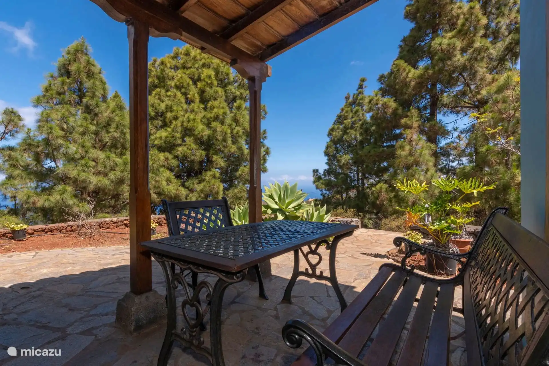 The terrace covered from the inside: wrought iron table and benches under the wooden beams, with the pine trees and the sea in the background.