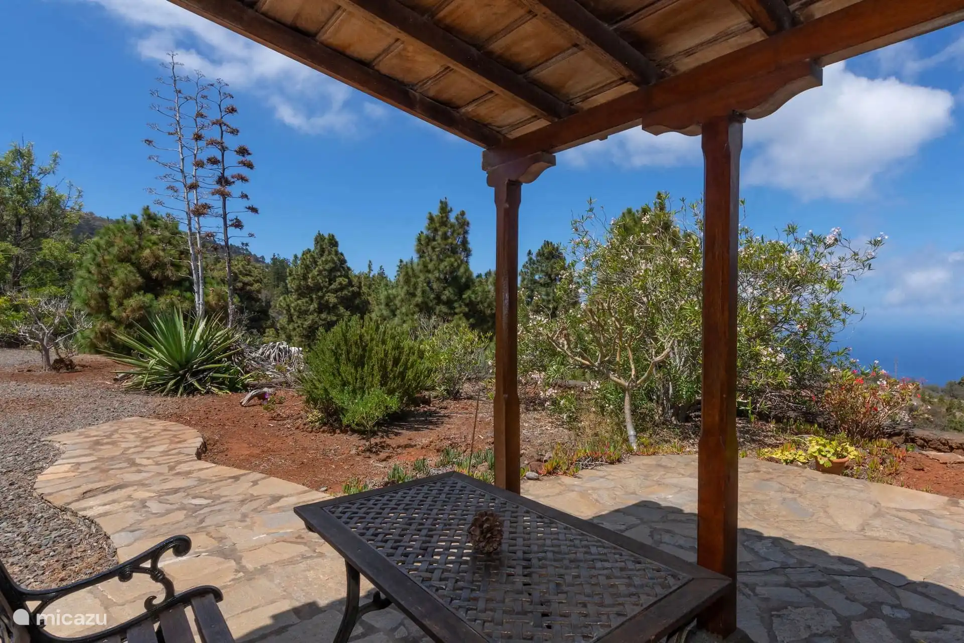 Überdachte Terrasse mit Schmiedeeisentisch und Bank auf Plattenboden; Vorne der Garten mit Oleandern und Kiefern, im Hintergrund das Meer.