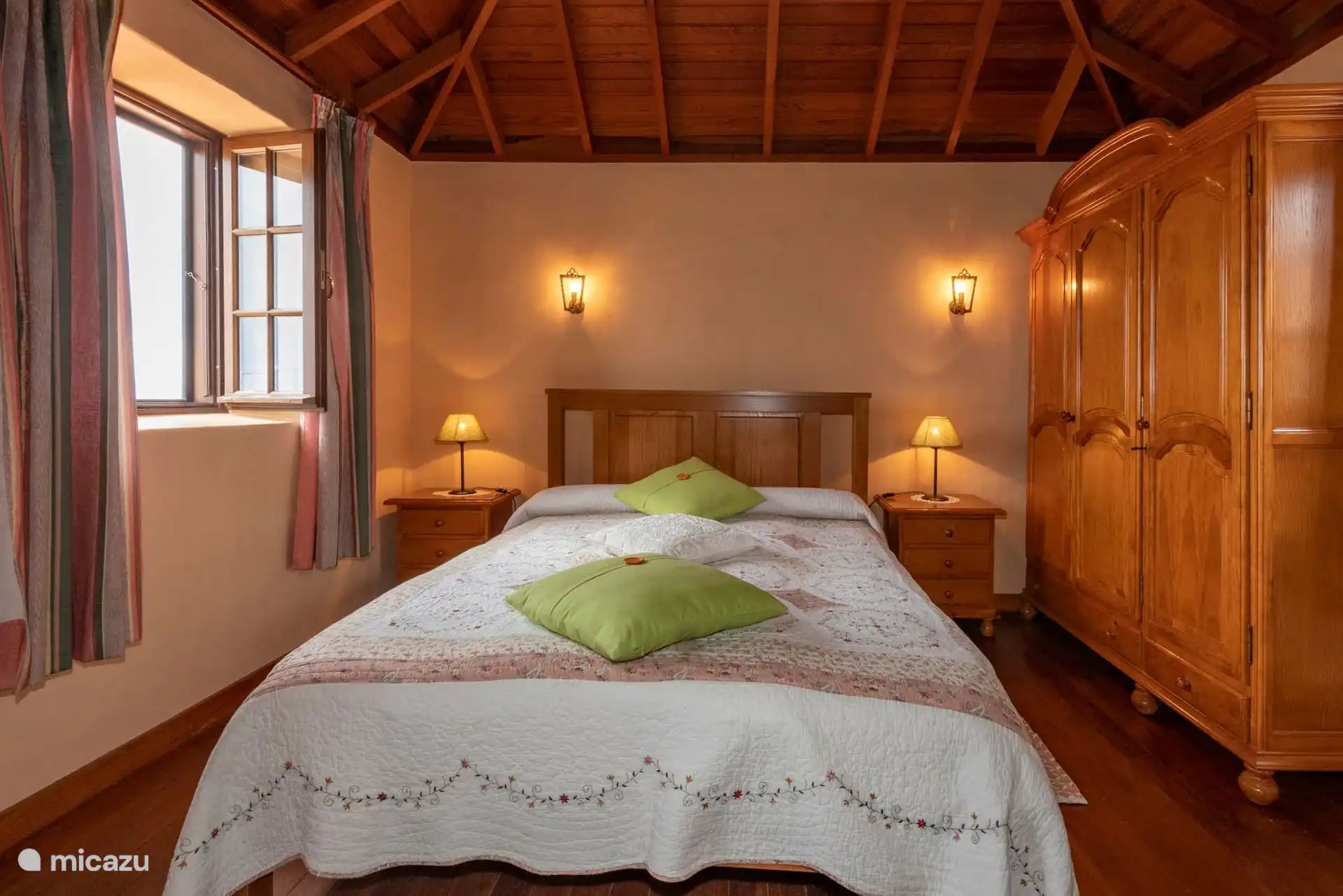 The same bedroom in front, with the sconces lit, the two bedside tables and the wardrobe next to the open window.