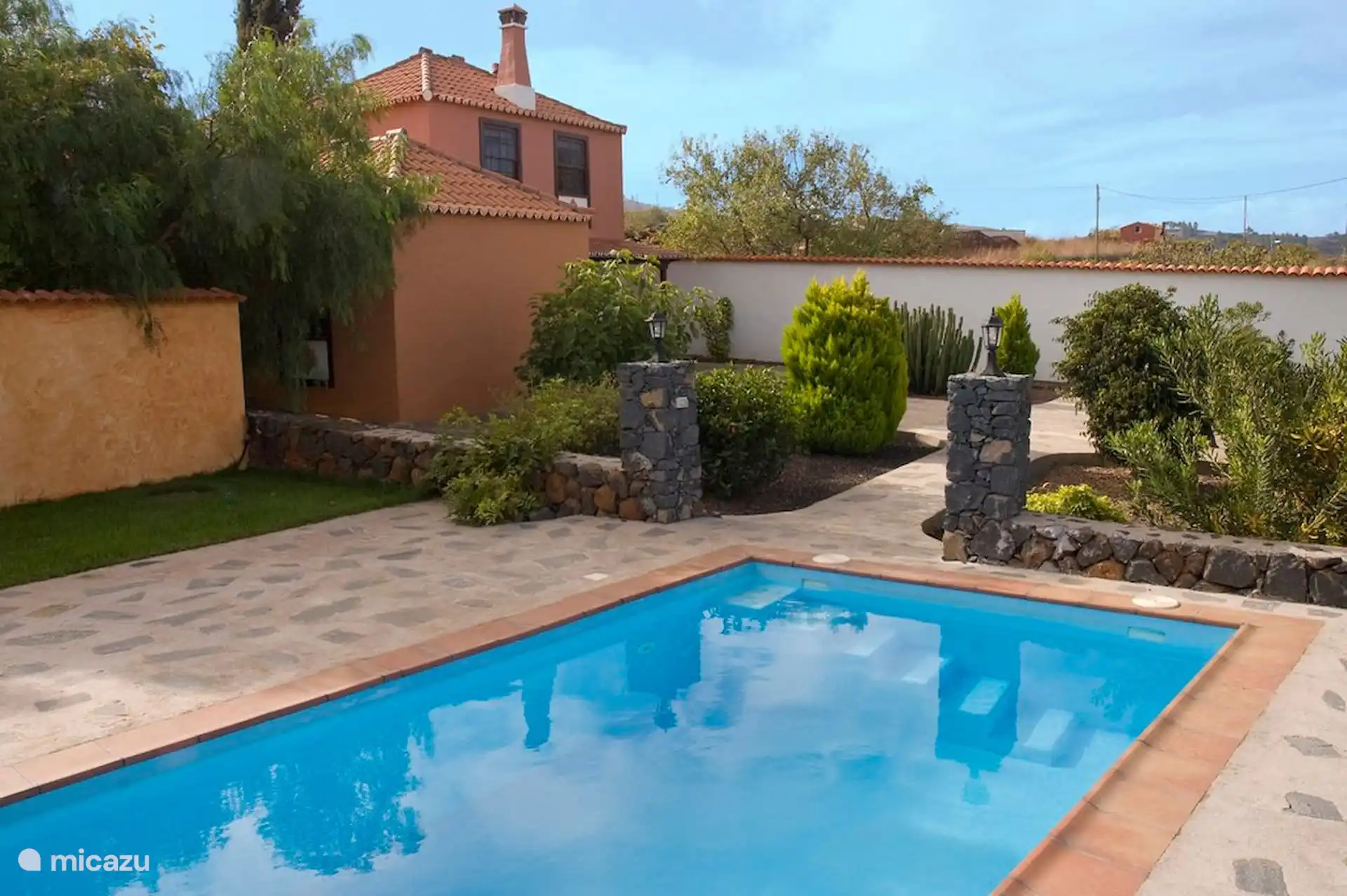 The swimming pool in the garden, with the two-storey house in the background; on the sides, pillars and walls of volcanic stone with lanterns.