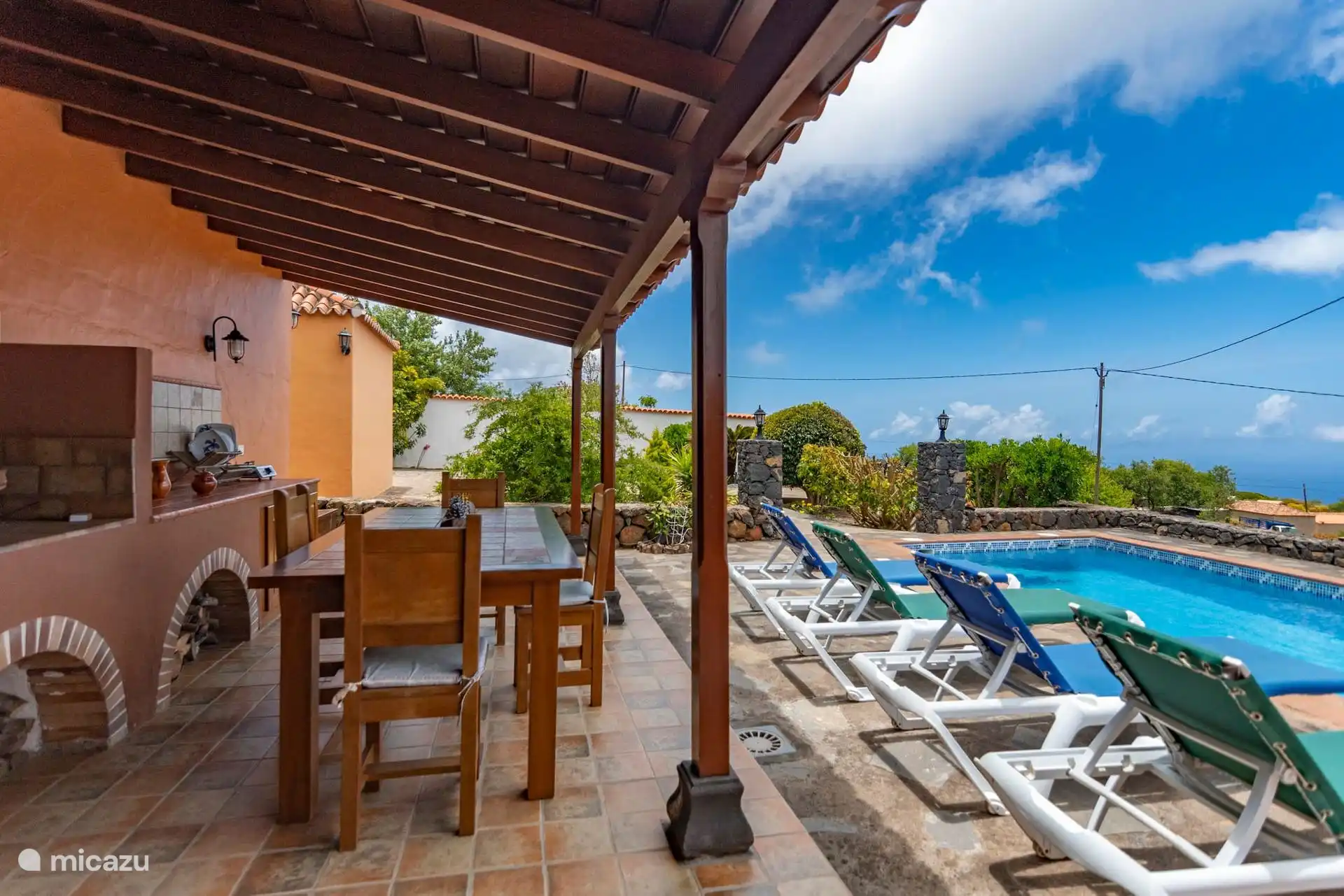 Outdoor dining area under the porch, next to the built-in barbecue; on the right, the sun loungers, the swimming pool and the sea in the background.