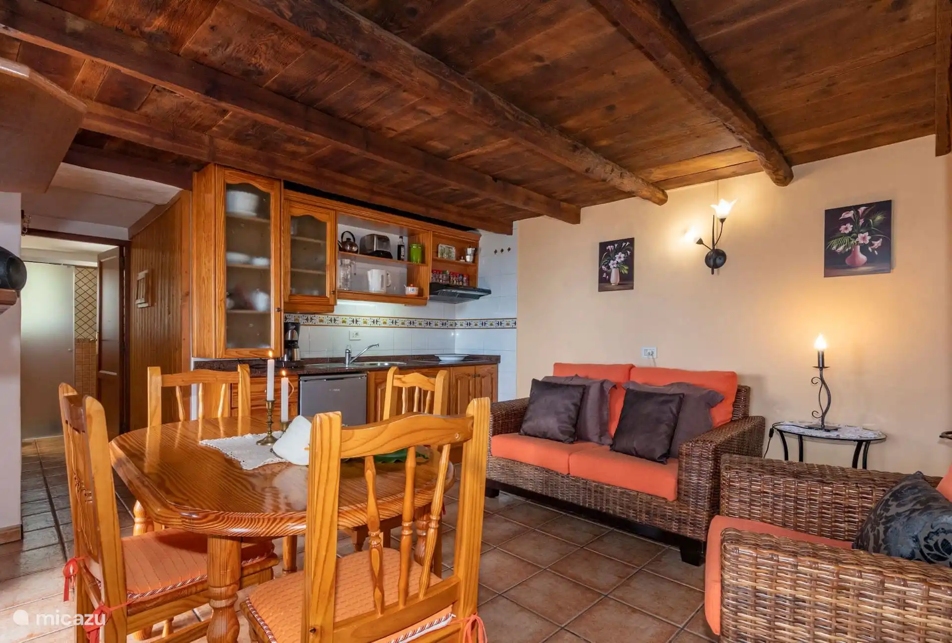 Living-dining room with integrated kitchen at the back, wooden table and wicker sofas, under the beamed ceiling.