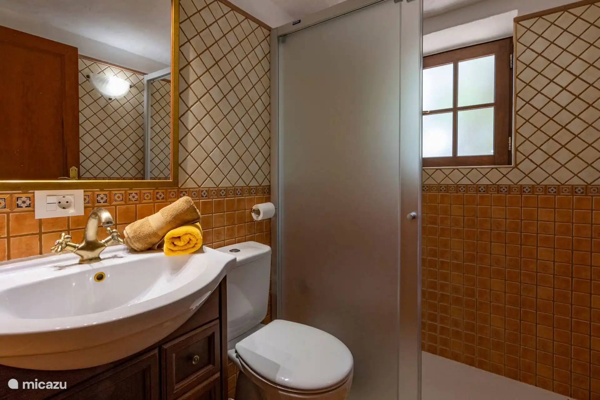Bathroom with sink on wooden furniture, toilet and shower with screen; Wooden window and two-tone tiling.