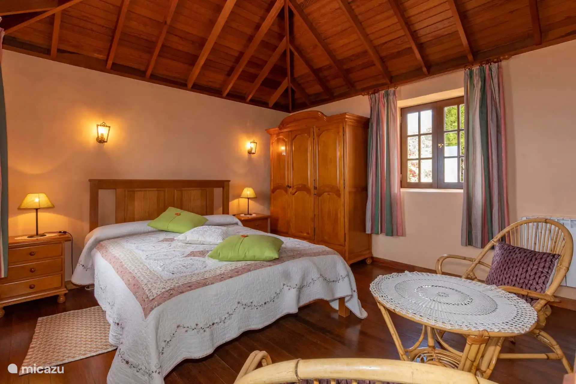 Bedroom with double bed under gabled wooden roof; wardrobe, bedside tables, wooden floor and a wicker corner by the window.