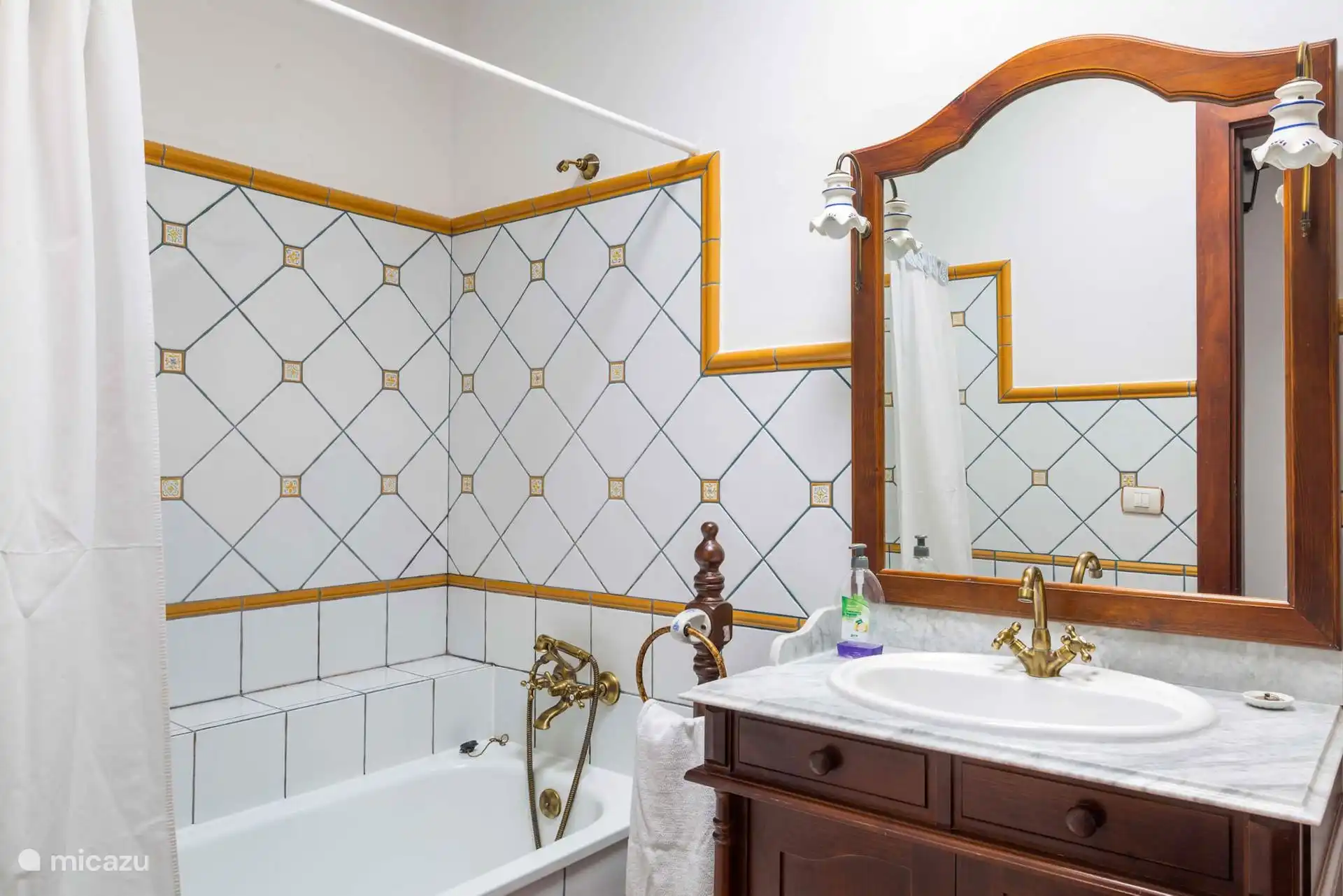 Badkamer met bad en gordijn, betegeld met gele rand; de wastafel, op een houten kast met een marmeren aanrechtblad.