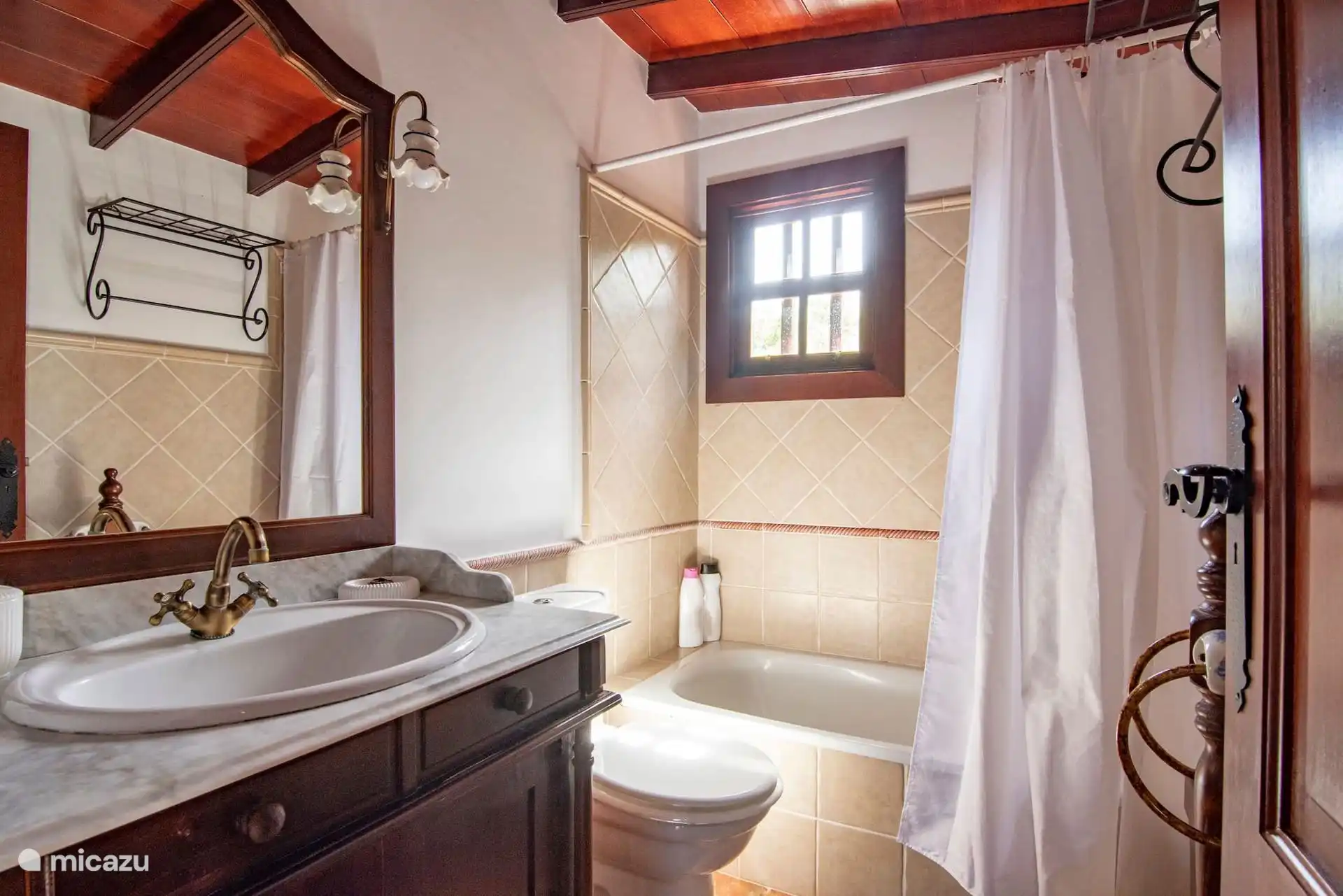 Badkamer met bad en gordijn, wastafel op een houten kast met marmeren aanrecht en houten raam.