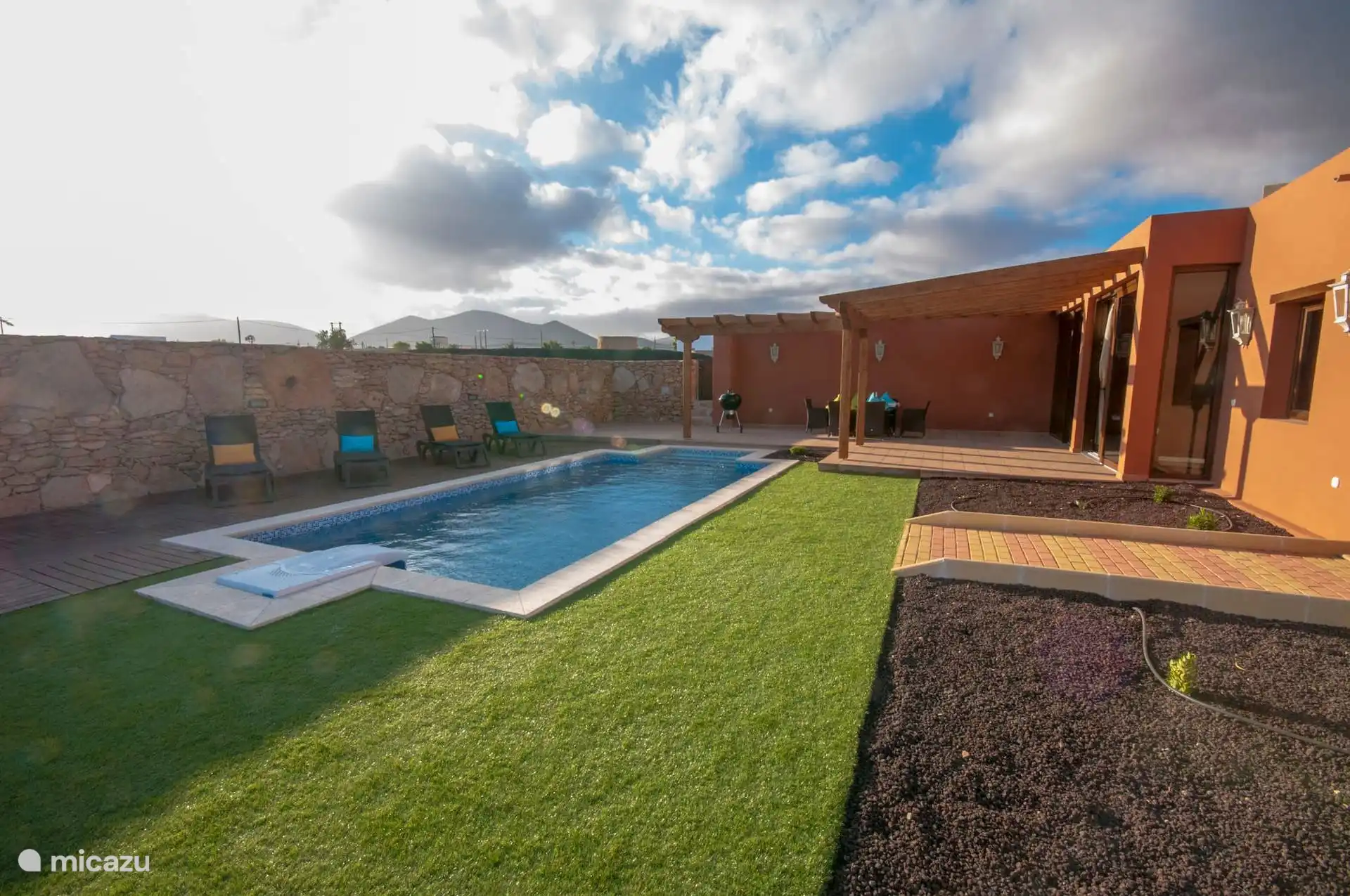 The lawned garden, swimming pool and sun loungers next to the stone wall; in the background, the bare mountains.