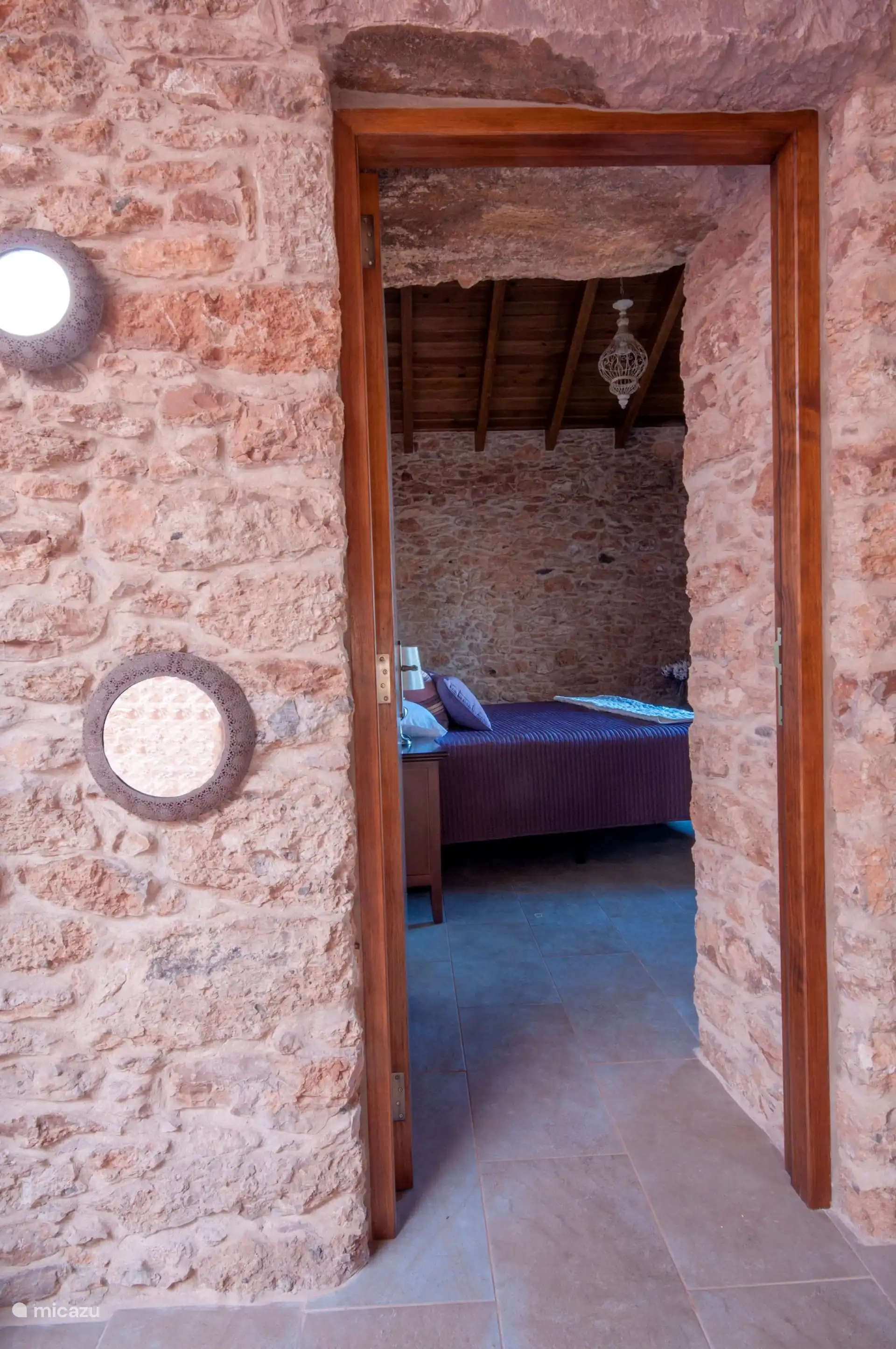 From the hallway, the wooden door opens into one of the bedrooms; on the stone wall, round mirrors.