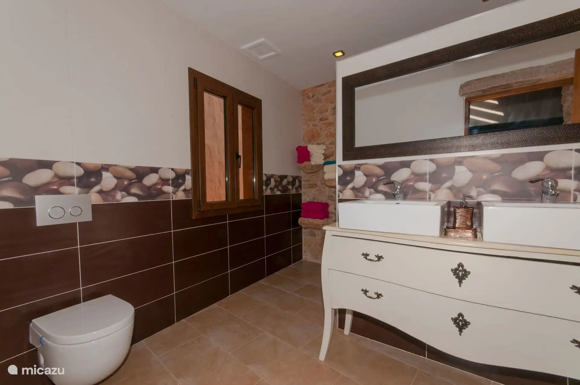 Spacious bathroom with two sinks on a chest of drawers, elongated mirror and wooden window; the towels, on the stone ledge.