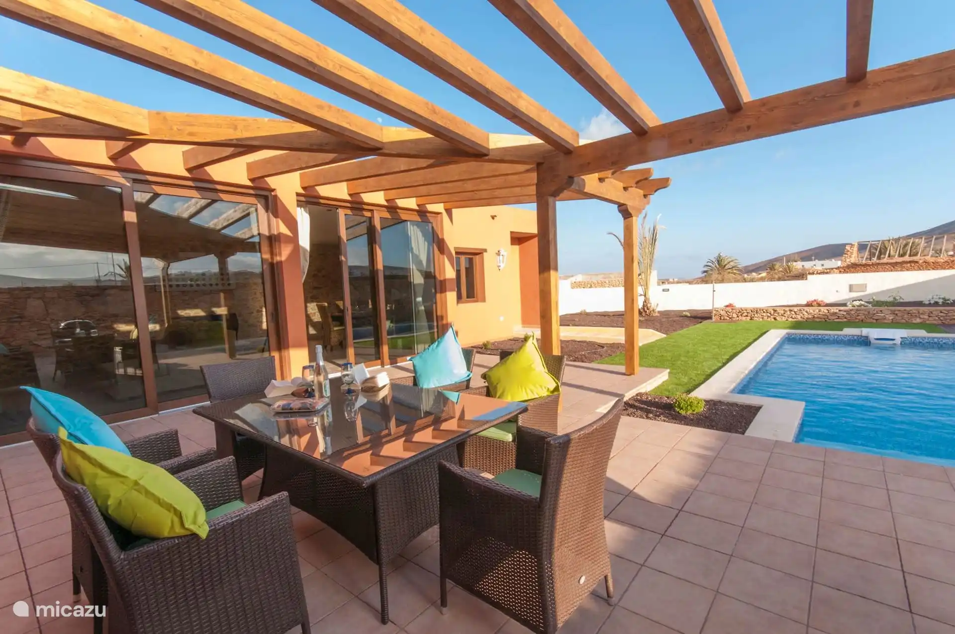 Terrace covered by the wooden pergola, with table and fibre armchairs next to the pool.