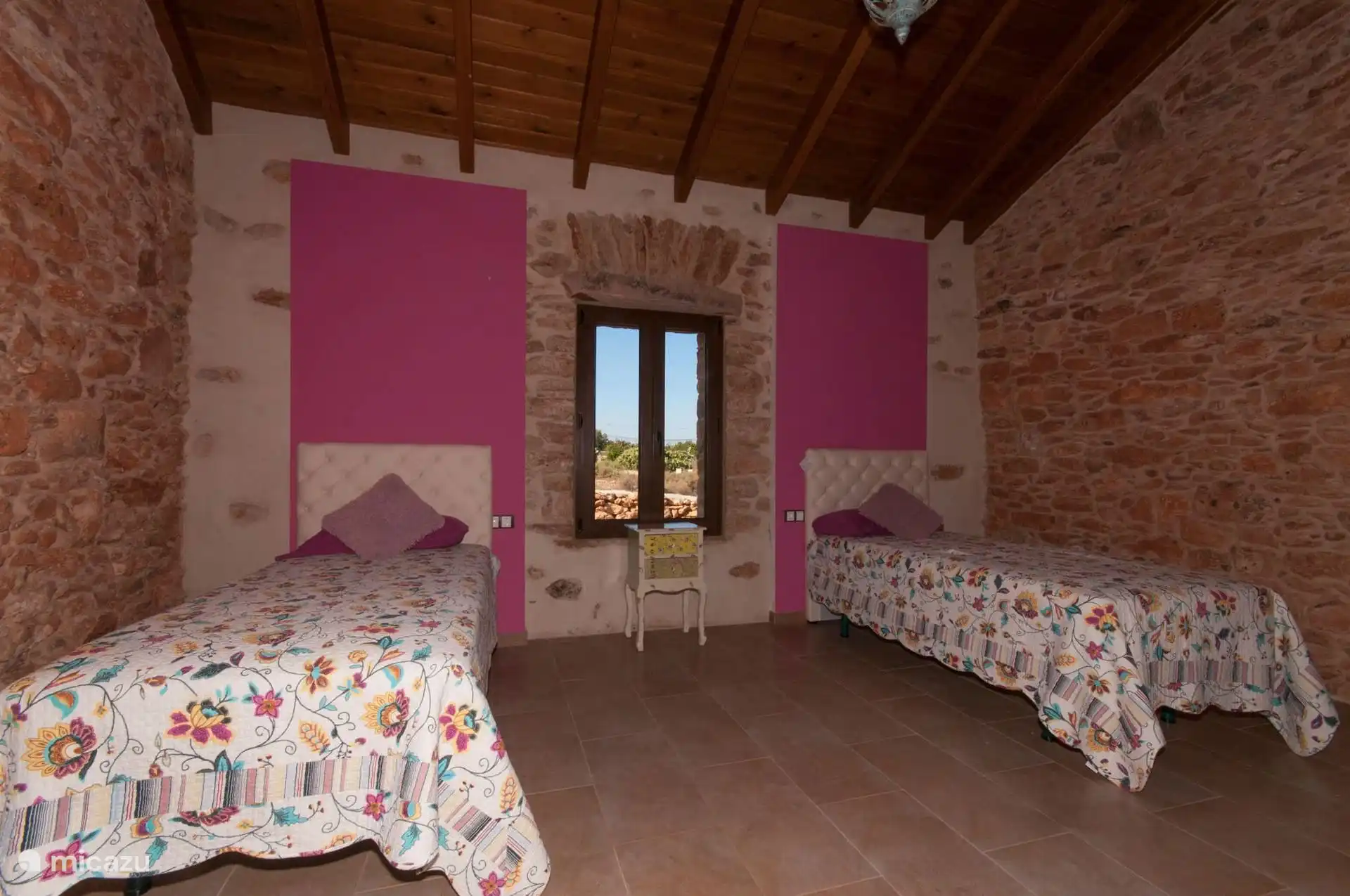 Bedroom with two single beds under the ceiling of beams and wooden board, between stone walls.
