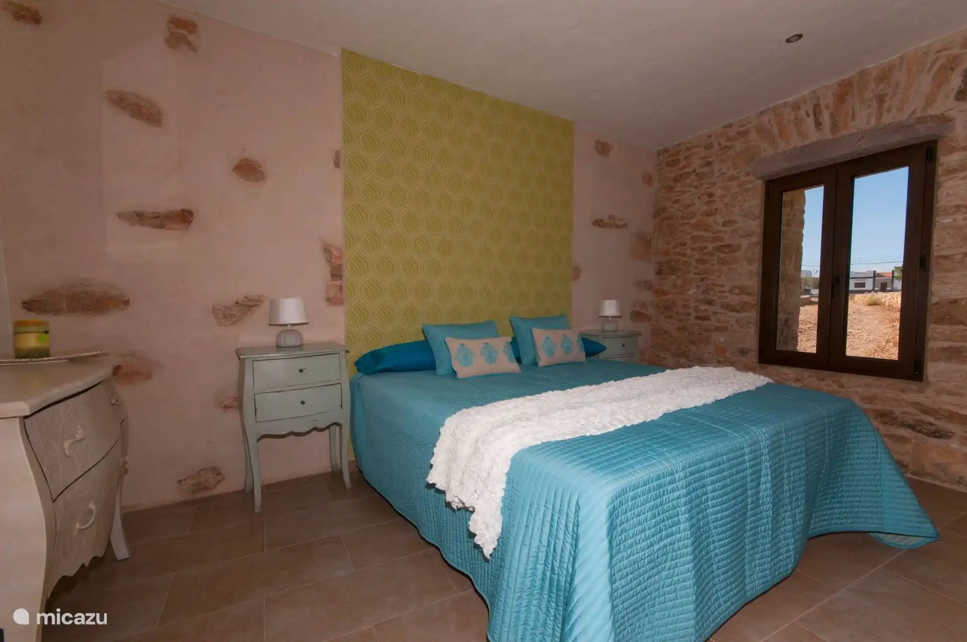 Bedroom with double bed between the stone wall and the wallpapered wall; The window has a stone frame.