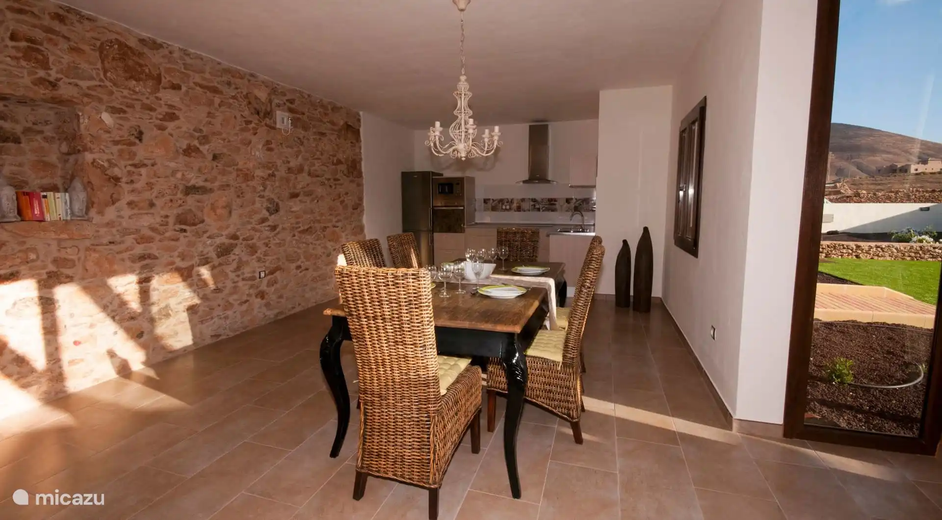 Dining room with wooden table and wicker chairs against the stone wall; at the back, the kitchen with oven, microwave and hood.