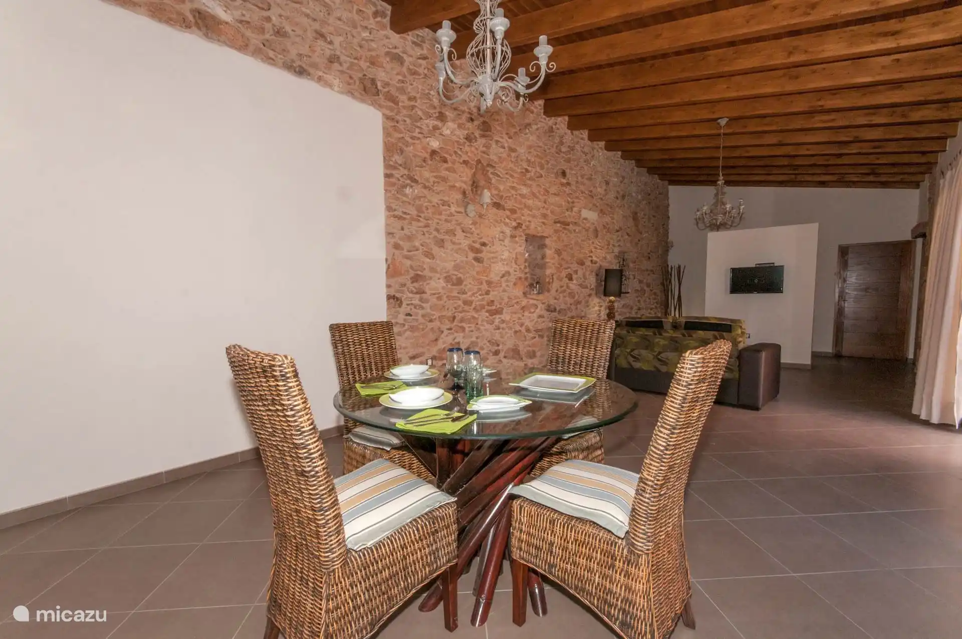 Eetkamer met ronde glazen tafel en vier rotanstoelen, met de tafel gedekt; Daarachter de loungeruimte.