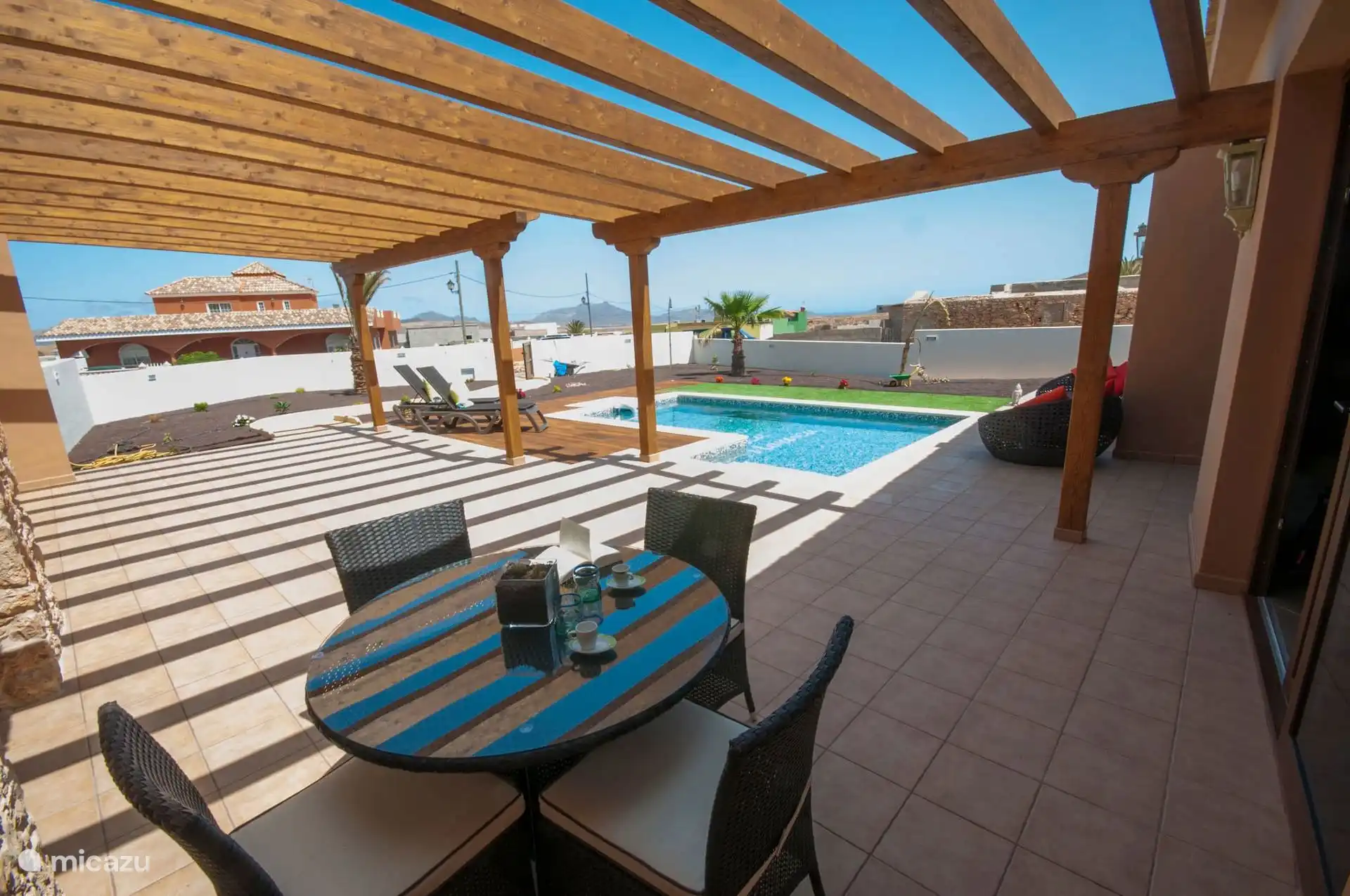 Terras overdekt door de pergola, met ronde tafel en vier stoelen; Voor de zaal, het zwembad en de ligstoelen.