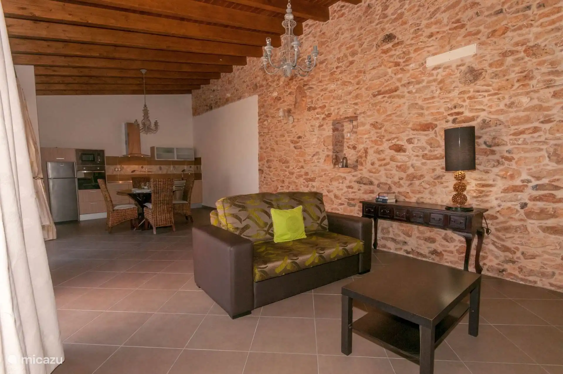 Woonkamer met bank en lage tafel onder het balkenplafond; achterin de keuken en de eettafel met rieten stoelen.
