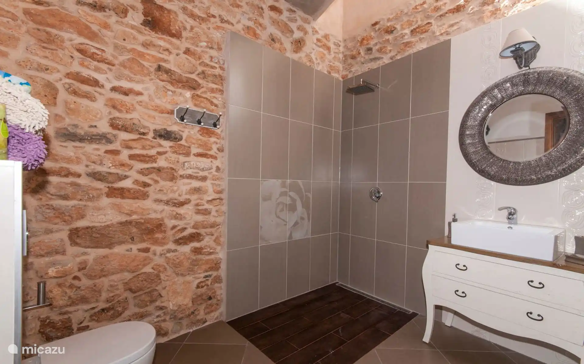 Badkamer met ingebouwde douche en douchekop, wasbak op een witte ladekast en ronde spiegel, naast de stenen muur.