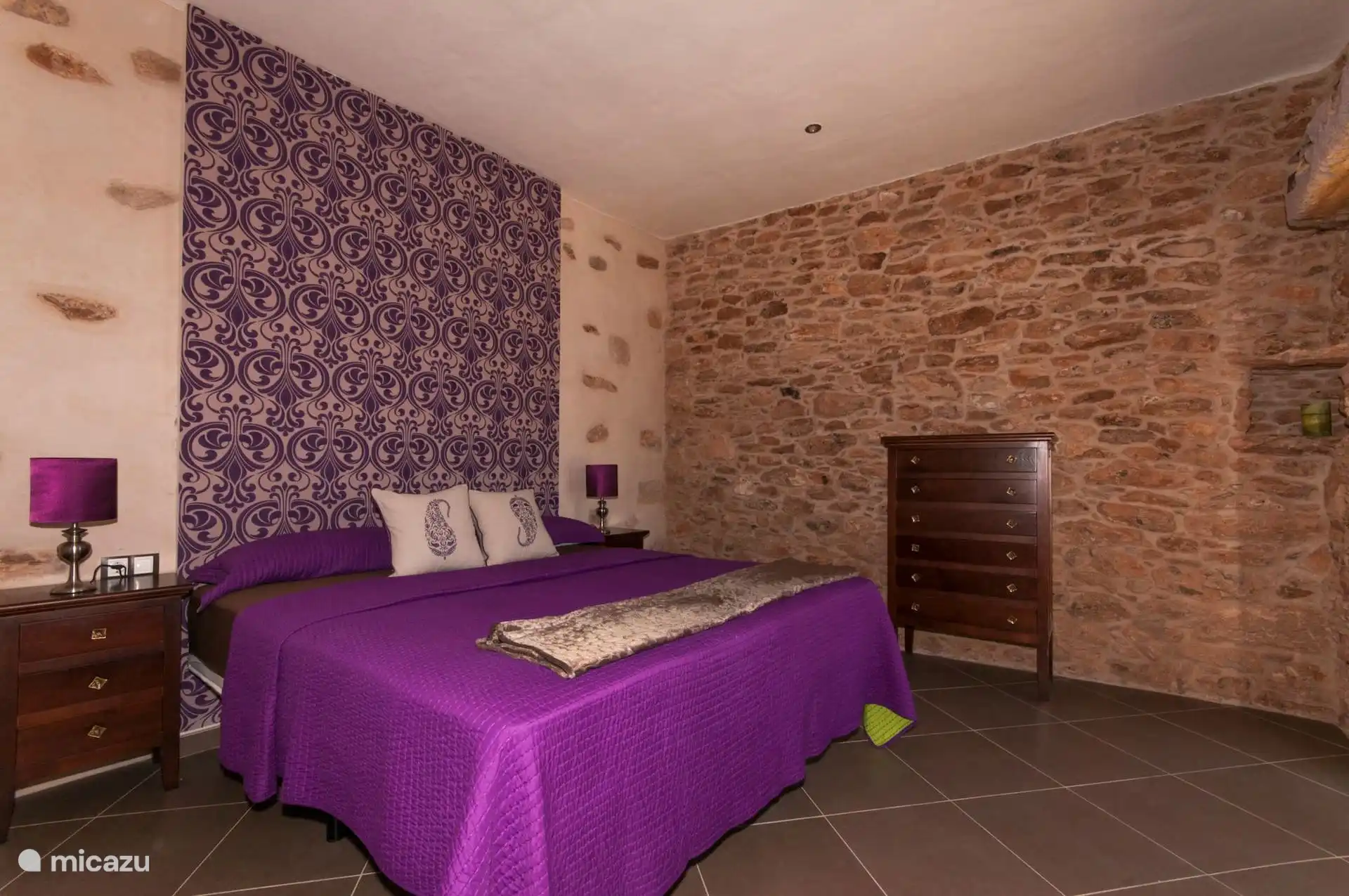 Slaapkamer met tweepersoonsbed, nachtkastjes en houten ladekast, tussen de blootliggende stenen muur en het behang.