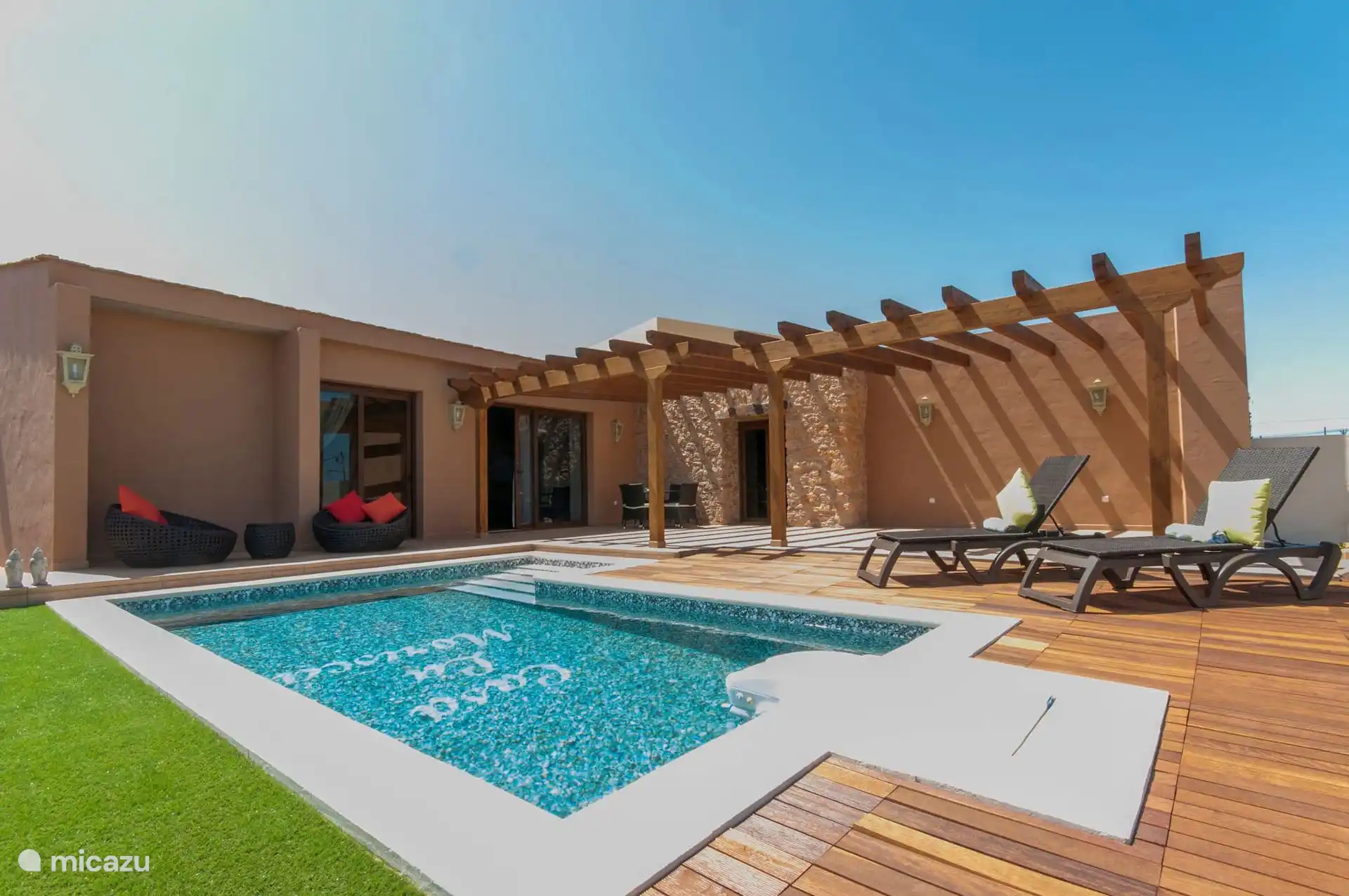 Het huis gezien vanaf het gazon: gevel met houten pergola, ligstoelen op het terras en het zwembad op de voorgrond.