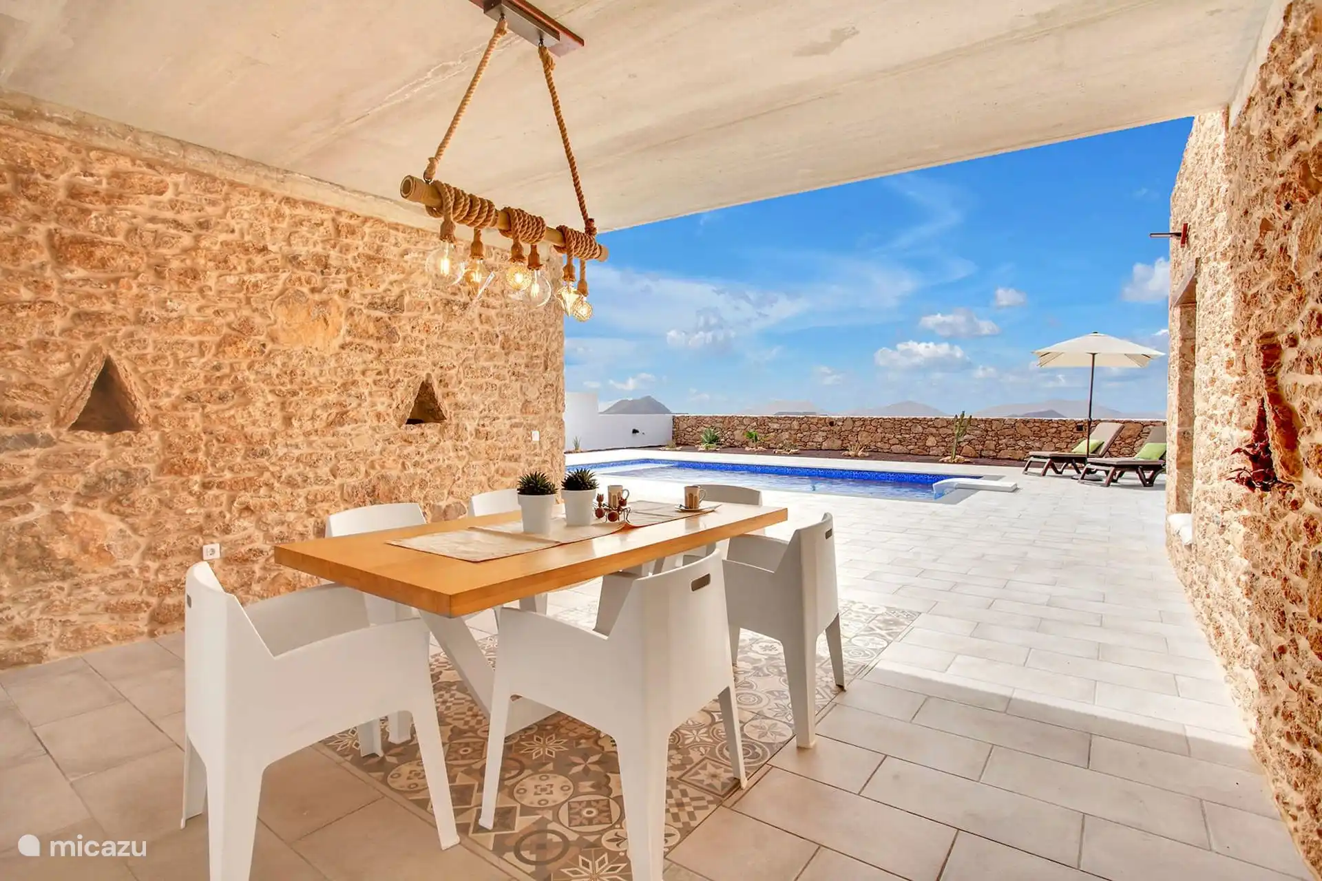 Porche cubierto con mesa de madera, sillas blancas y lámpara de cuerda con bombillas; delante, la piscina.