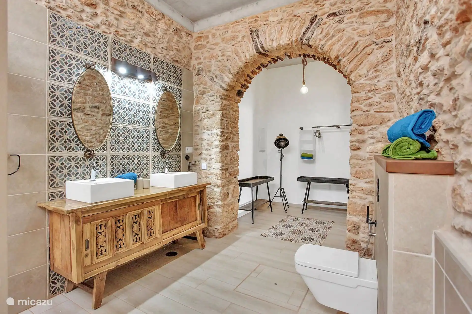 Baño con doble lavabo sobre mueble de madera tallada, espejos ovalados y arco de piedra hacia la zona del inodoro.