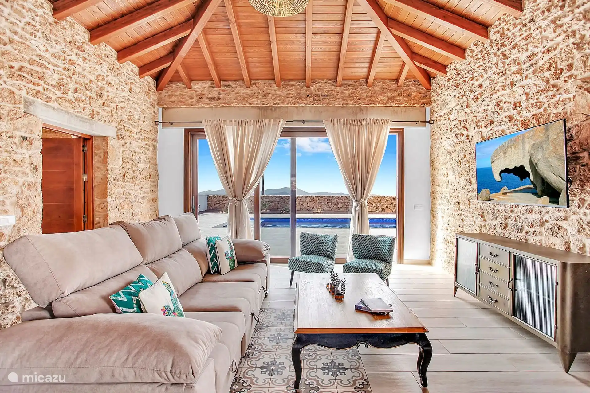 Salón con sofá esquinero, dos butacas y televisión sobre el muro de piedra, bajo el techo de vigas de madera; la cristalera da a la piscina.
