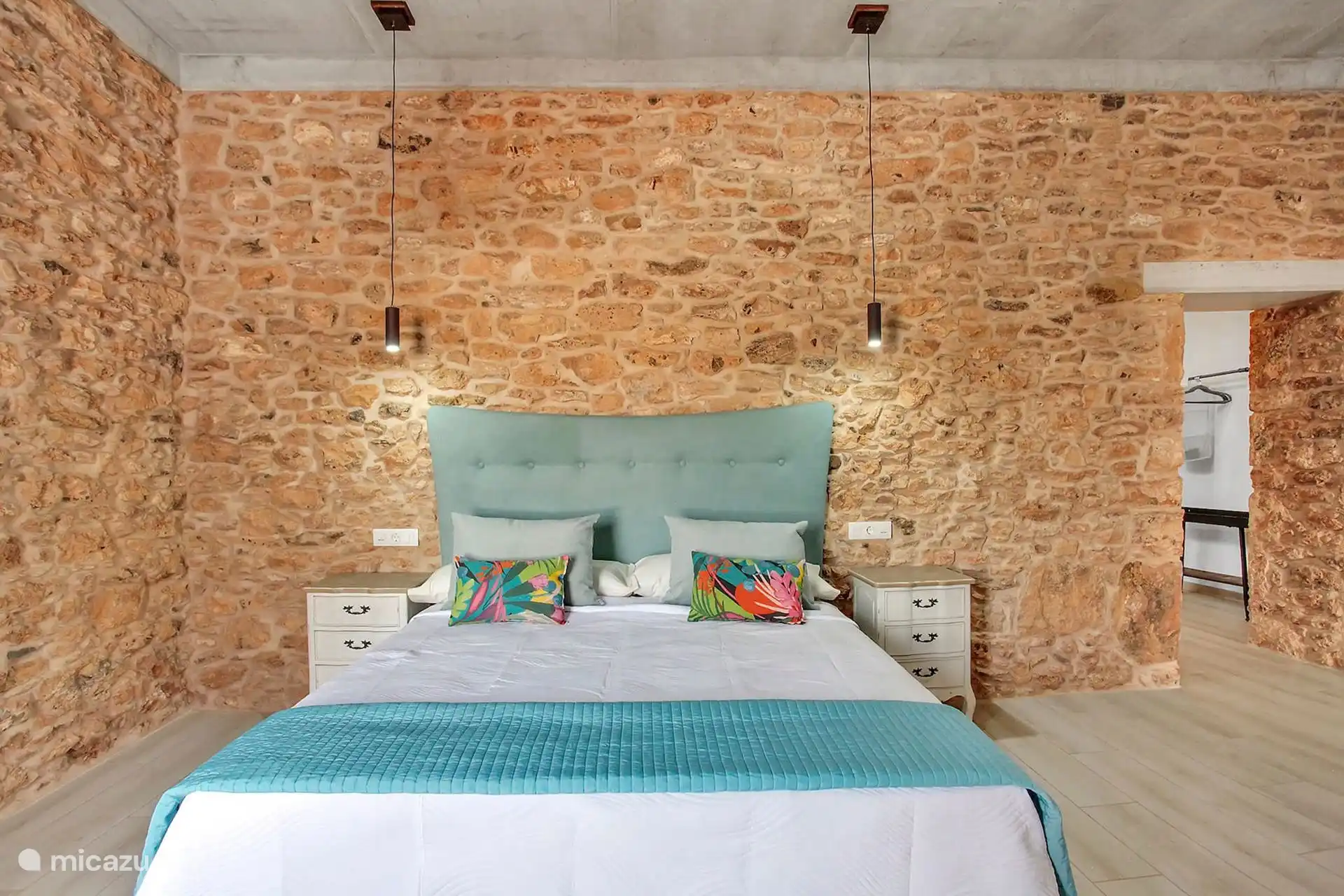 Dormitorio con cabecero turquesa, mesillas blancas y dos lámparas colgantes sobre la pared de piedra.