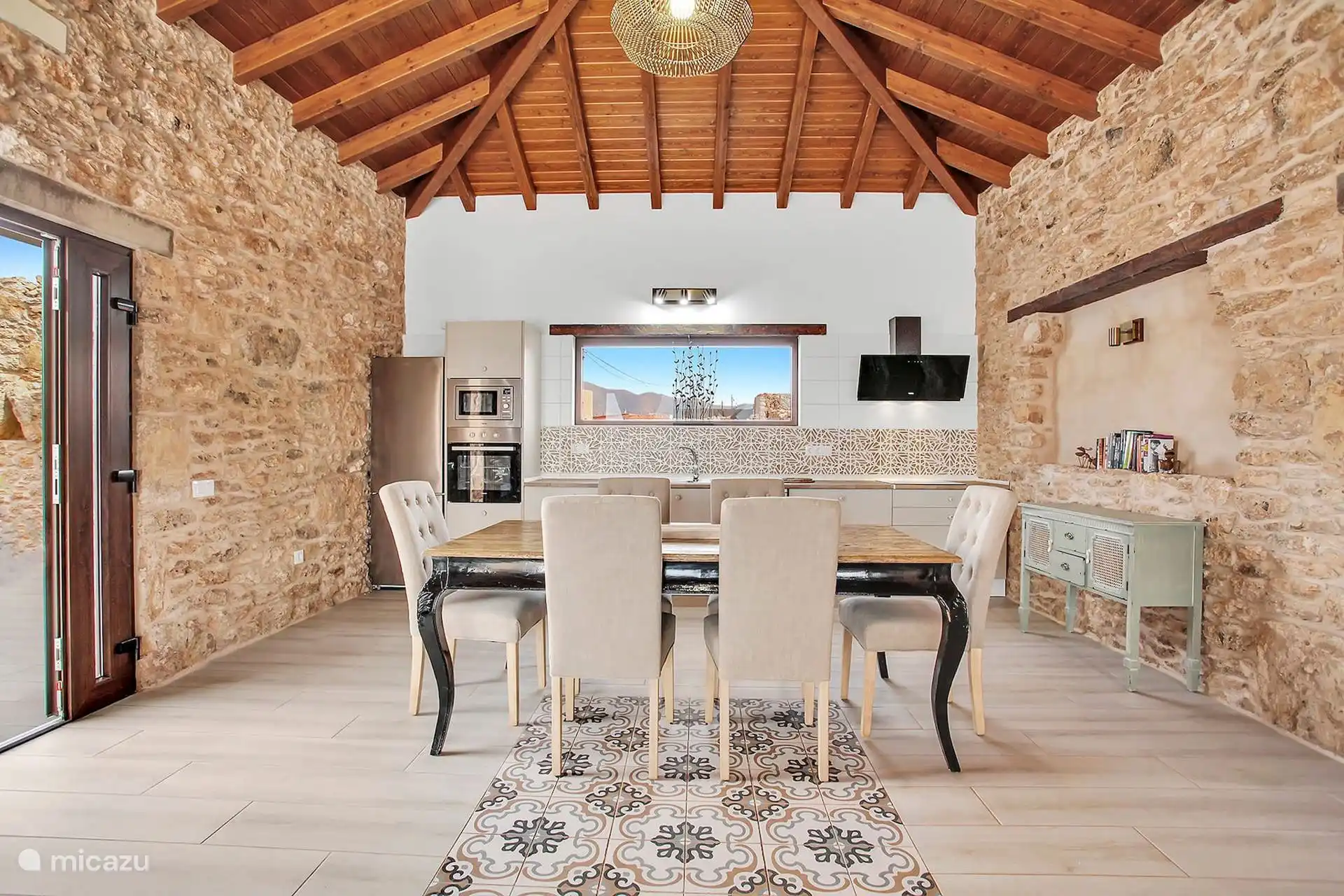 Comedor para seis con la cocina al fondo: horno, microondas, nevera y una ventana alargada con las montañas.