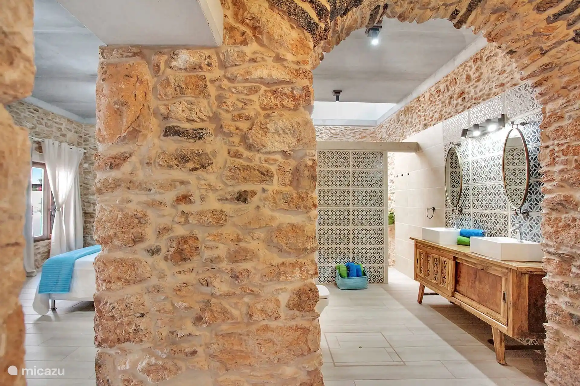 Baño abierto al dormitorio, separado por el muro de piedra; mueble de madera con dos lavabos y celosía al fondo.