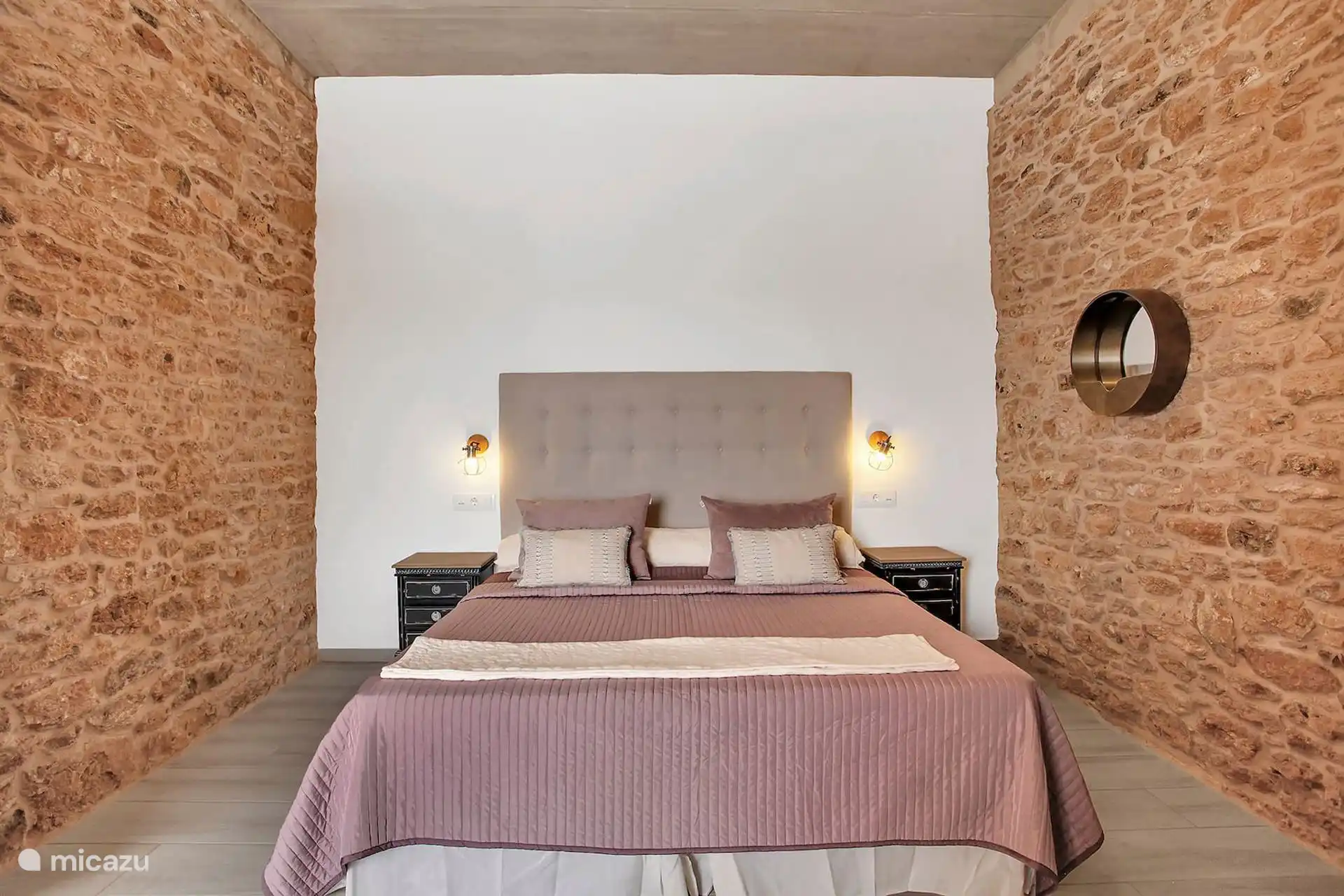 Dormitorio con cama de matrimonio, cabecero tapizado y apliques a ambos lados, entre paredes de piedra vista.
