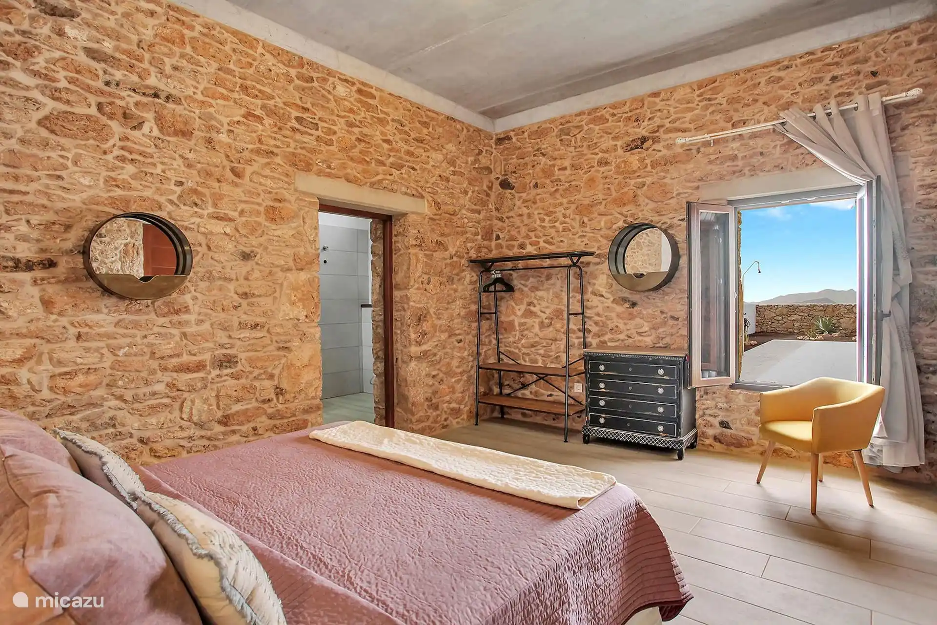 Otra vista del dormitorio, con burro para la ropa, cómoda oscura y ventana abierta a la terraza y a las montañas.