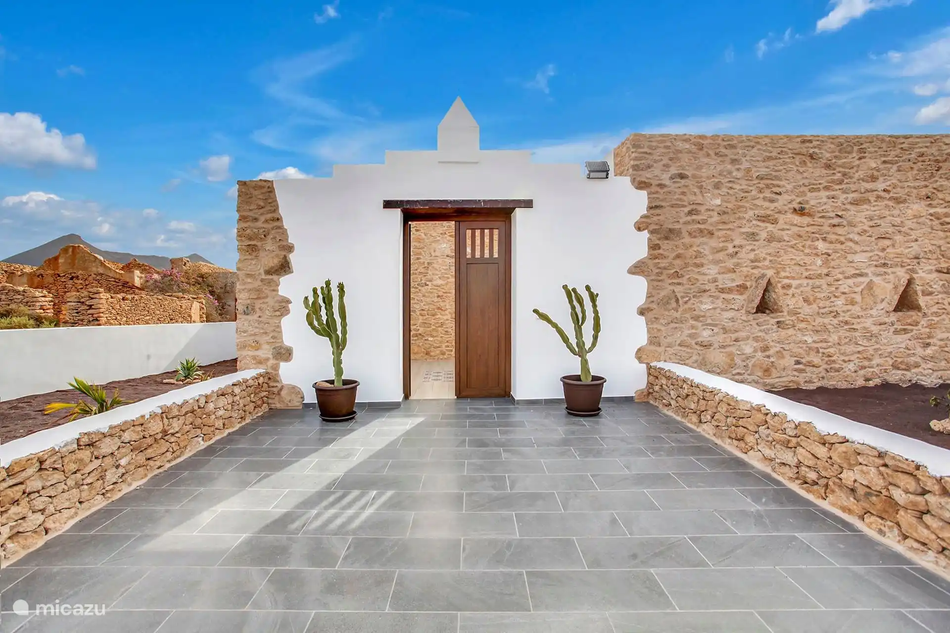 Entrada de la casa: fachada encalada con puerta de madera, dos cactus en maceta y muretes de piedra seca.