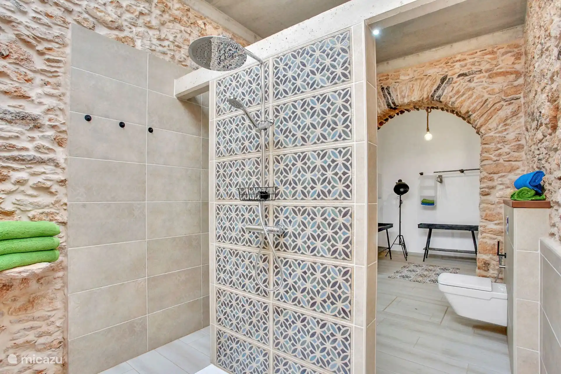Ducha con rociador y frente de azulejo estampado; al otro lado del arco, el inodoro y las toallas.