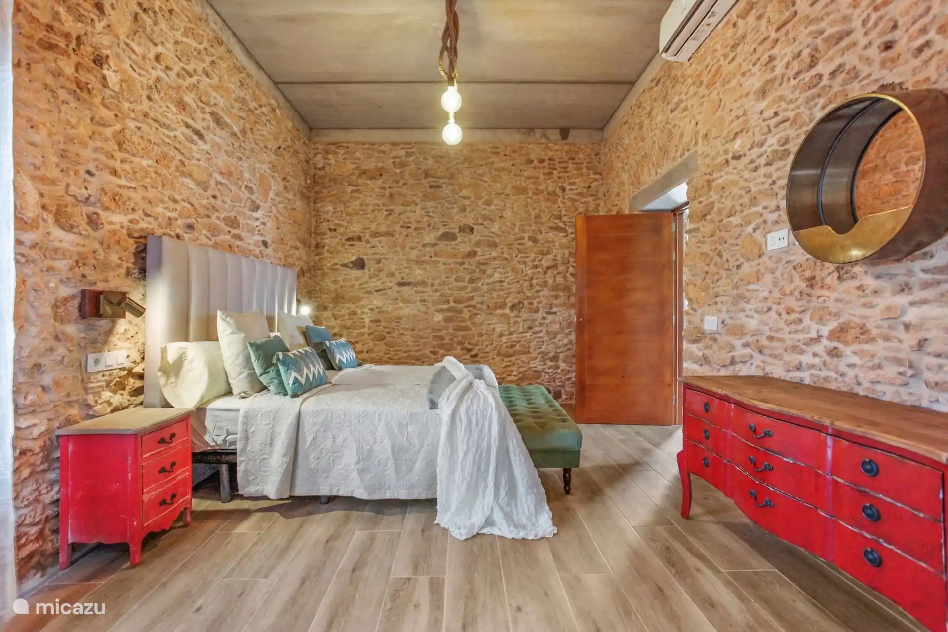 Dezelfde slaapkamer vanaf het voeteneind van het bed, met de rode ladekast, de ronde spiegel en de airconditioning op de steen.