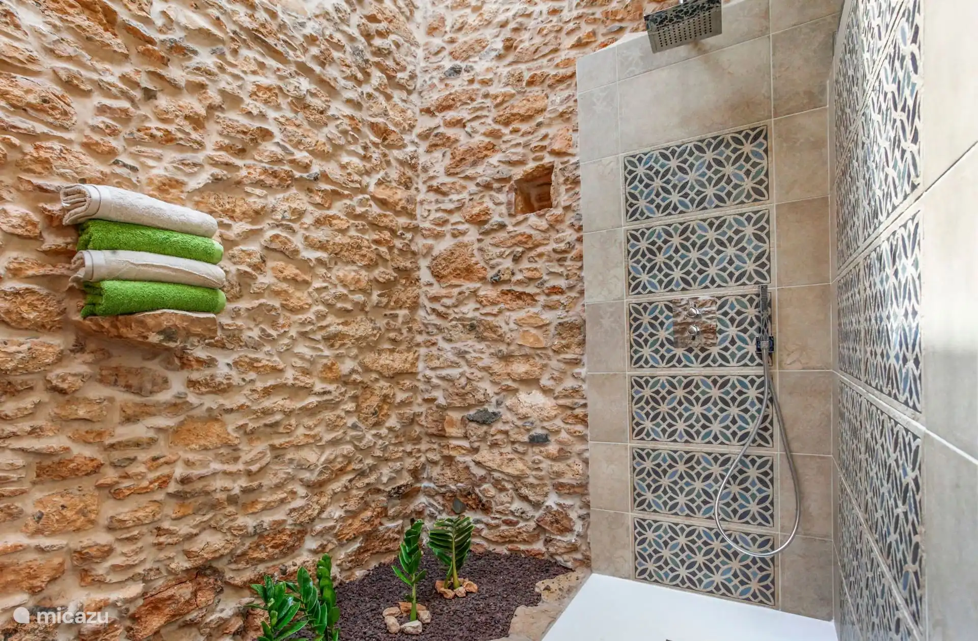 Details van de douche, met de regendouchekop, de handdoeken op de stenen richel en een plant op het bed van picón.