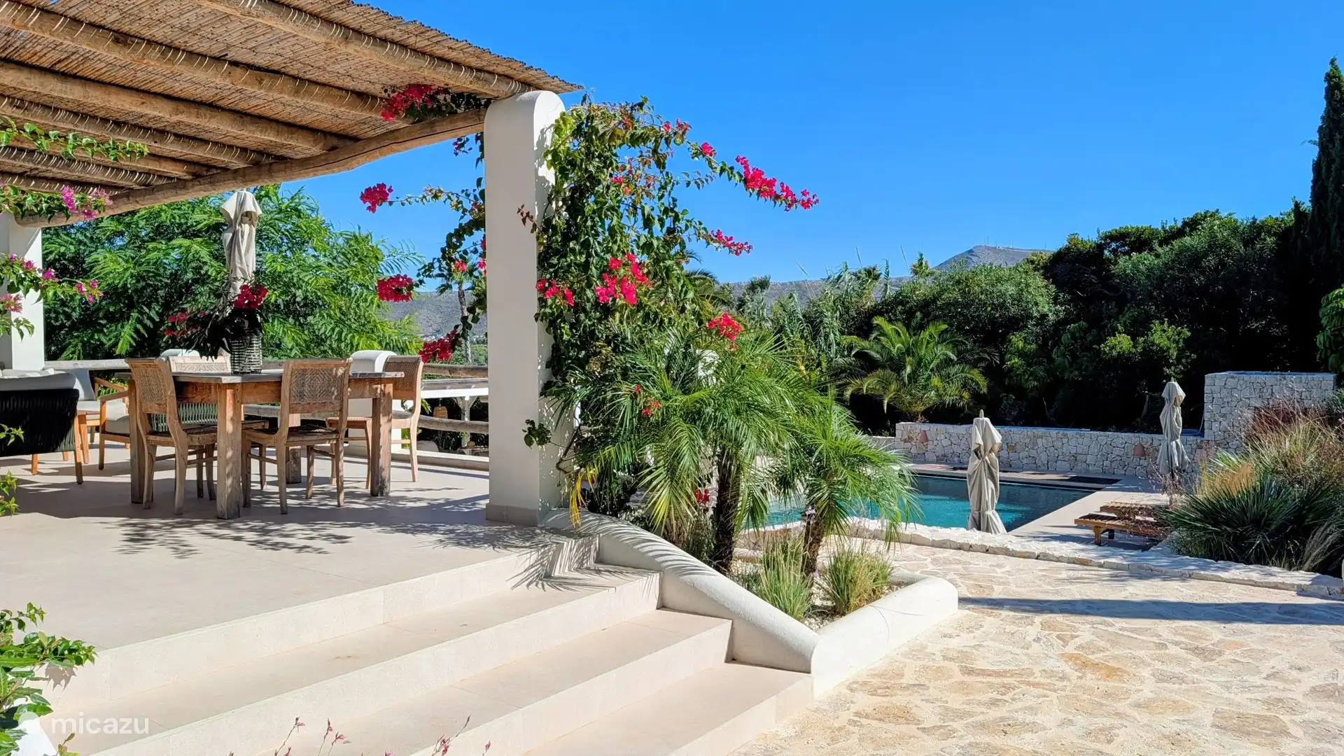 Terras onder de pergola met eettafel en bloeiende bougainville, uitkijkend op het zwembad. Zo geniet je van je lunch met zicht op het water.