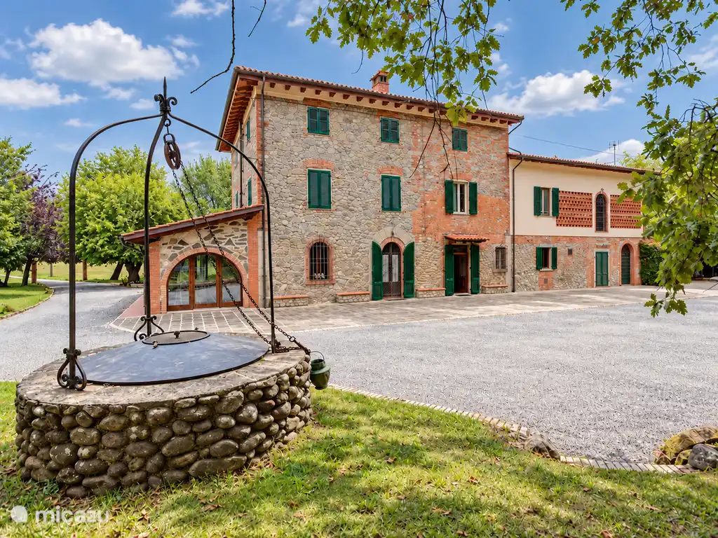 Vue extérieure de la maison en pierre, avec un puits traditionnel au premier plan et une verdure entourant la propriété.