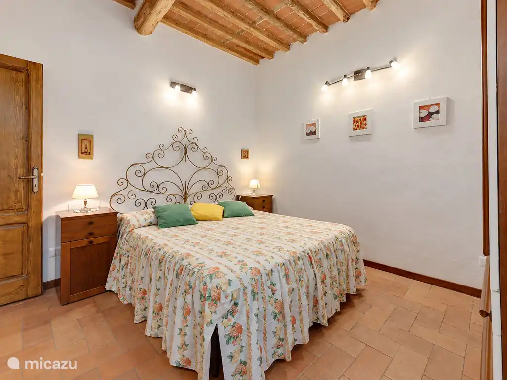 Chambre double avec une tête de lit en fer forgé, plafond à poutres en bois et sol en carreaux de terre cuite.