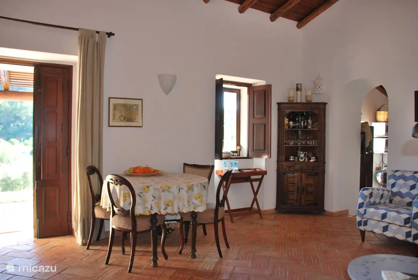 Otra vista del salón con la entrada a la cocina a la izquierda y la estufa de leña a la derecha. Hay contraventanas de madera delante de puertas y ventanas, con las que se puede mantener fuera el calor (y en los meses de invierno el frío).