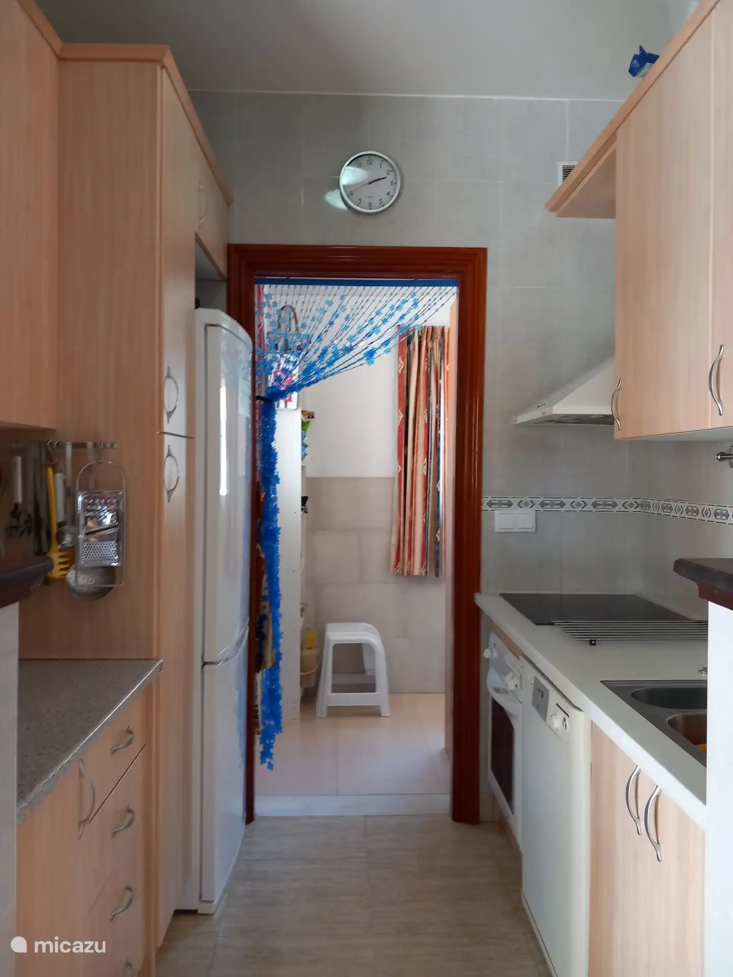 Doorkijk van keuken naar bijkeuken. De keuken heeft 2 werkbladen, dubbele spoelbak, keramische kookplaat, oven, afzuigkap, koelviescombinatie, koffiezetapparaat en vaatwasser.